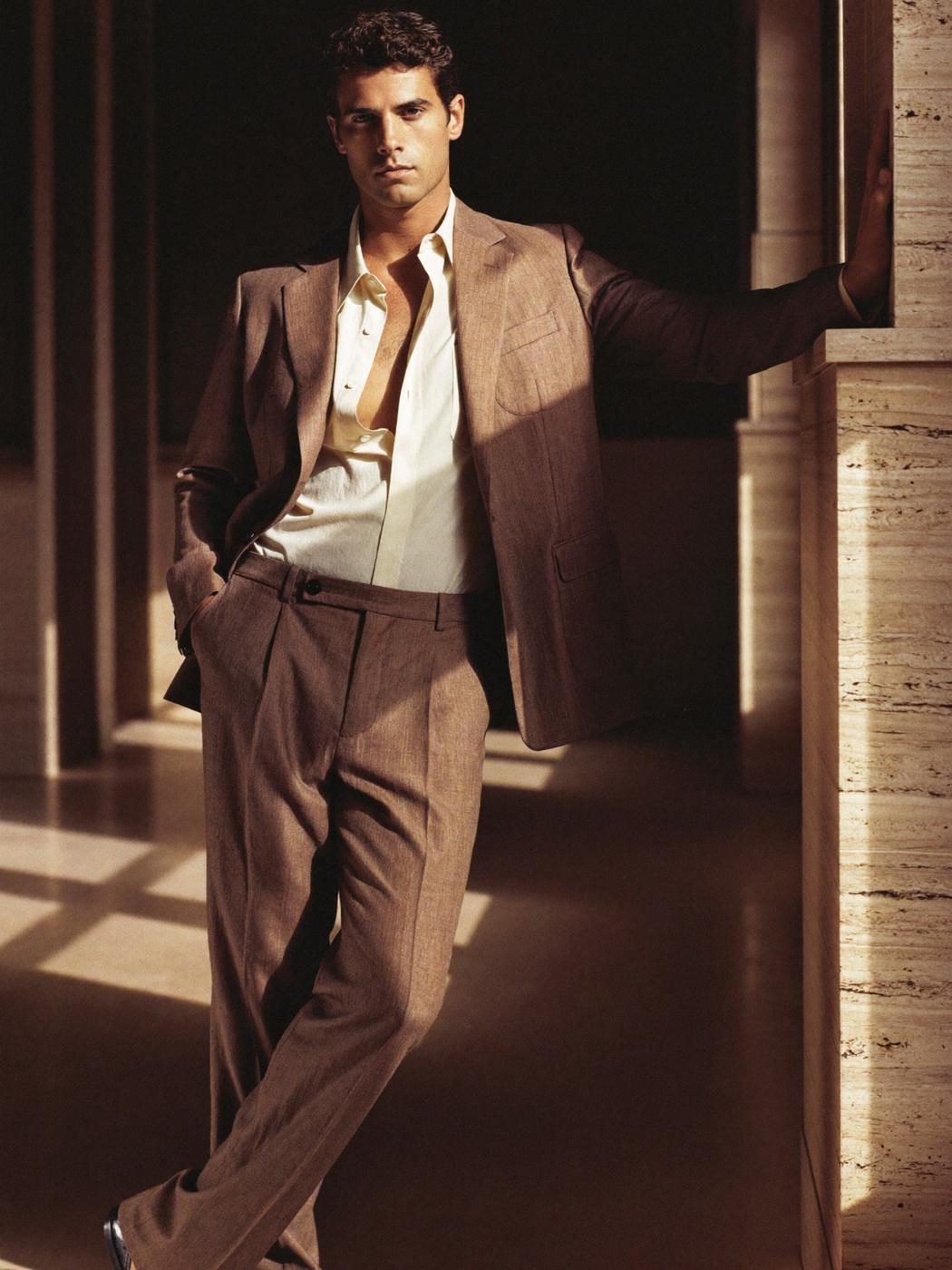 Man in a light brown linen suit and open-collared cream shirt, leaning against a travertine wall in a sunlit hallway