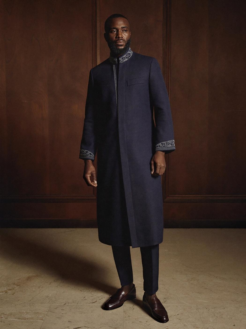 Man in a long midnight-navy Nigerian kaftan with embroidered collar and cuffs, in a walnut-panelled room