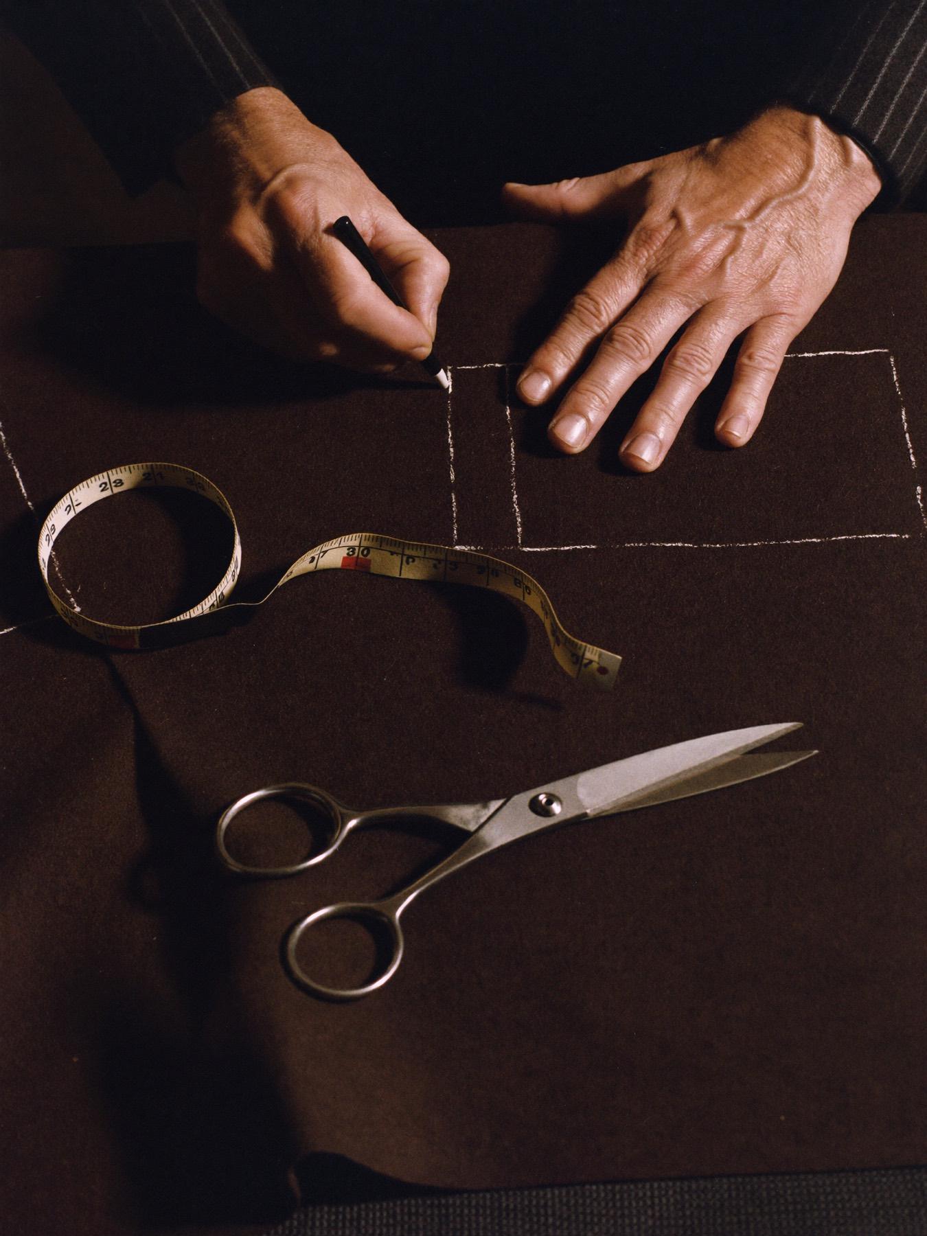 A tailor's hands chalking a pattern onto brown wool, beside a tape measure and shears