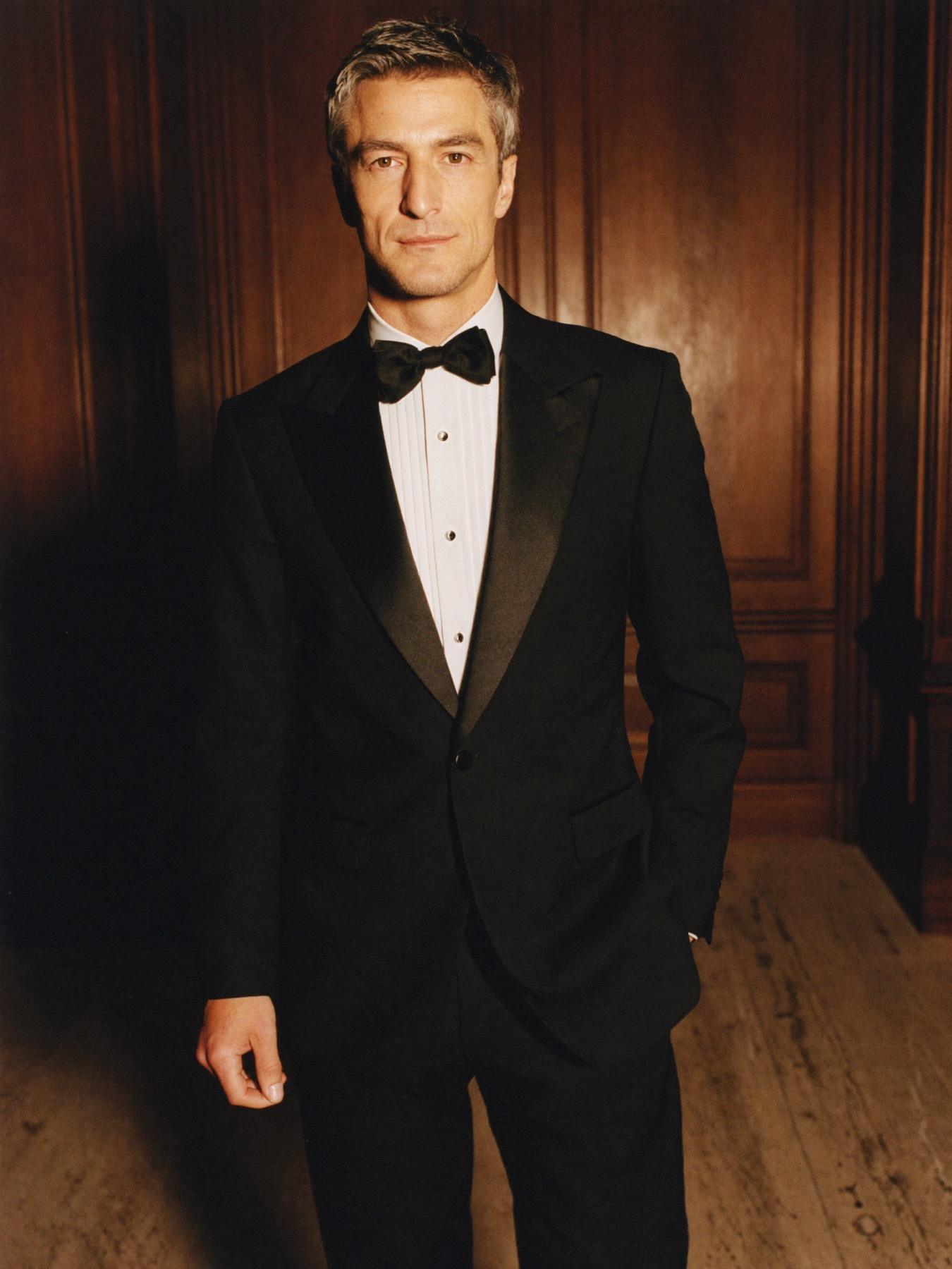 A man in a midnight peak-lapel tuxedo and silk bow tie in a walnut-panelled hall
